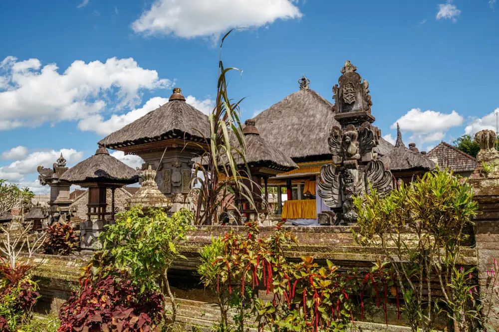 The traditional Balinese village of Penglipuran, showing its unique architecture with thatched roofs, ornate stone walls and gateways, and lush tropical plants under a blue sky.