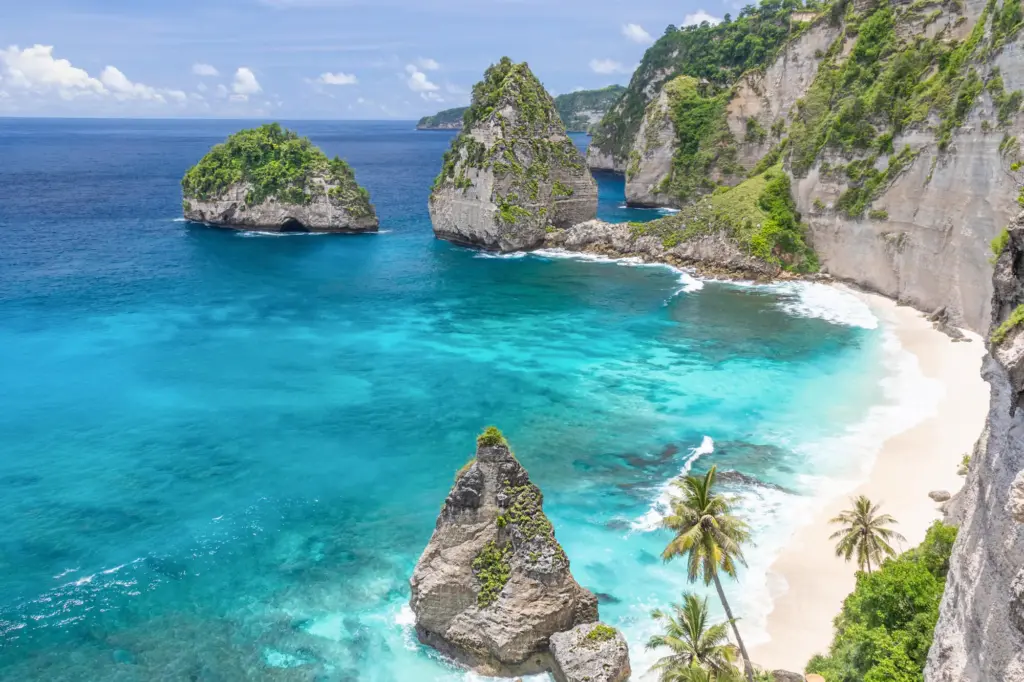 A high-angle view of Diamond Beach on Nusa Penida's east coast, showing the white sand beach nestled beneath limestone cliffs, vibrant turquoise water, and iconic pointed sea stacks.