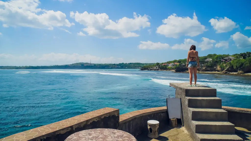 A woman in a bikini and shorts stands on a set of concrete steps on a clifftop, looking out at the wide, blue ocean and coastline on a sunny day in Bali.